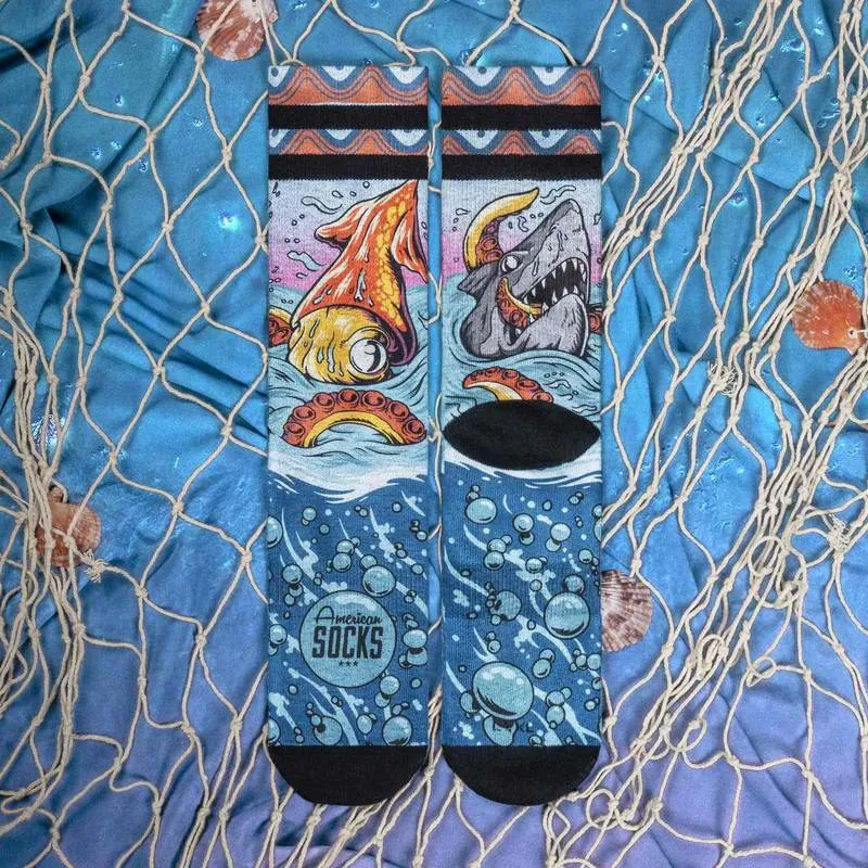 Deux chaussettes 'Seamonsters Mid High' avec pieuvre, requin, bulles et logo 'American Socks' sur tissu bleu avec coquillages et filet de pêche.
