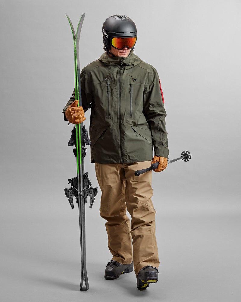 Skifahrer in GORE-TEX Jacke Skifahrer mit Z-2 HD GORE-TEX Pro 3L Shell Jacket Forest Green, Ski, Stöcken und Helm vor grauem Hintergrund.