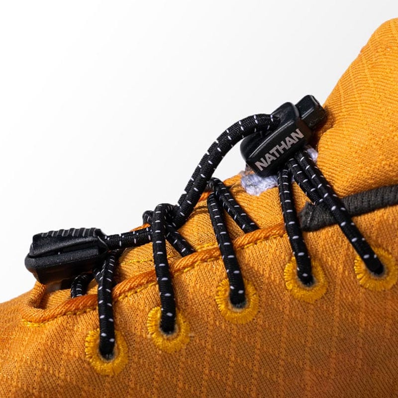 Gelber Schuh mit Nathan-Logo Detailansicht eines gelben Schuhs mit schwarzen Run Laces, die mit einem schwarzen Verschluss mit dem "Nathan"-Logo befestigt sind.