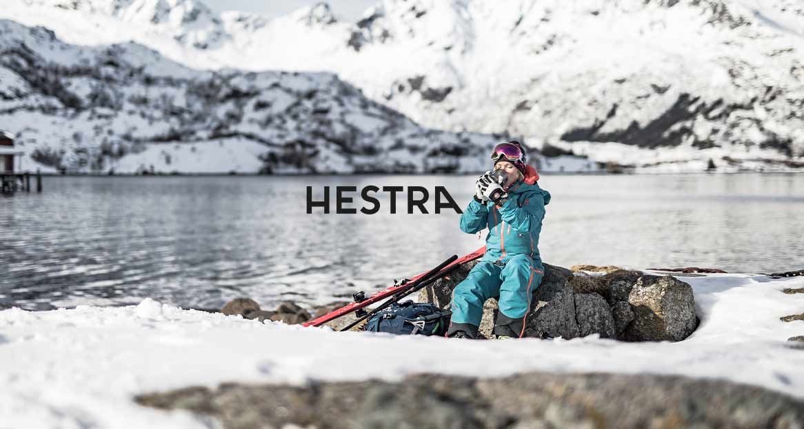 Woman in ski clothes drinking hot drink, sitting on rocks on the lakeshore with skis and mountains in the background.
