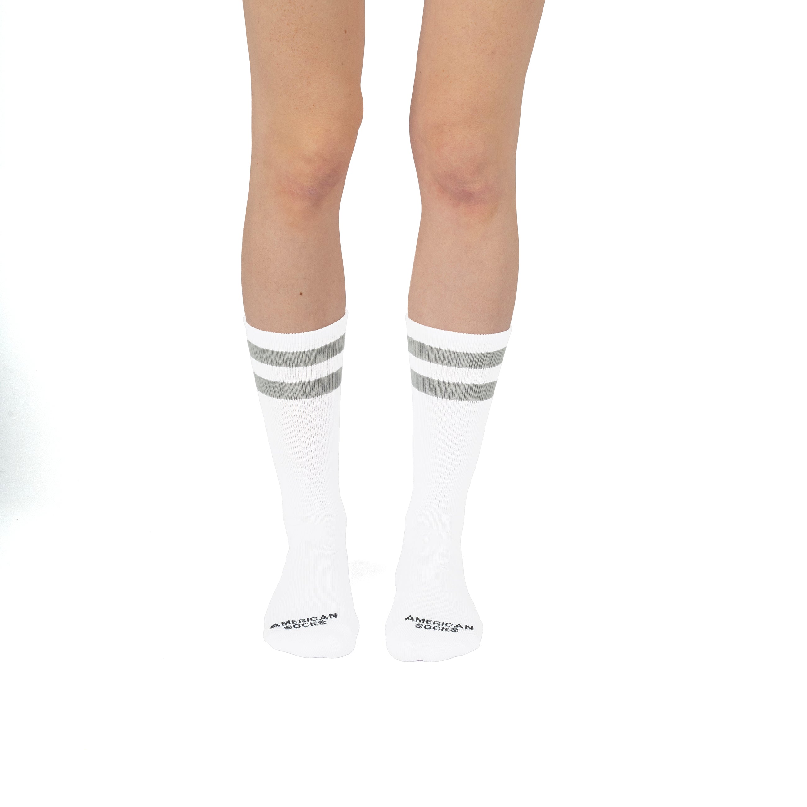 American Socks mit Streifen Beine mit weißen, mittelhohen Socken, zwei graue Streifen und Schriftzug "American Socks" auf den Füßen.