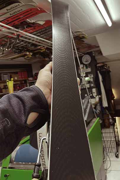 Ski mit Wellenmuster Eine Hand hält einen Ski hoch, der eine dunkle Basis mit einem sich wiederholenden Wellenmuster hat. Im Hintergrund sieht man Werkstatteinrichtung.