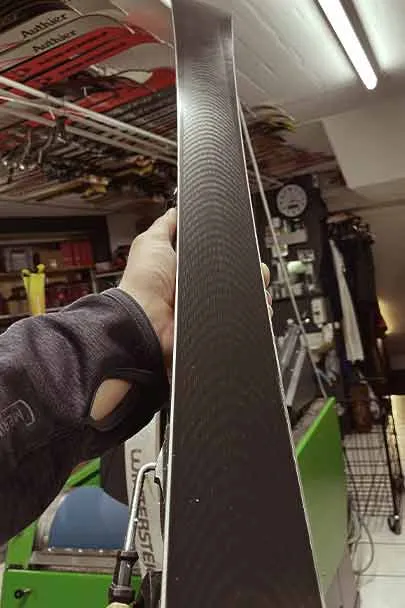 A hand holds up a ski, which has a dark base with a repeating wave pattern. Workshop equipment can be seen in the background.