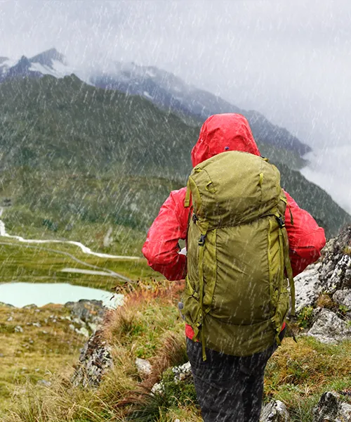 Wanderer in roter Jacke mit Rucksack im Regen vor Berglandschaft mit See.