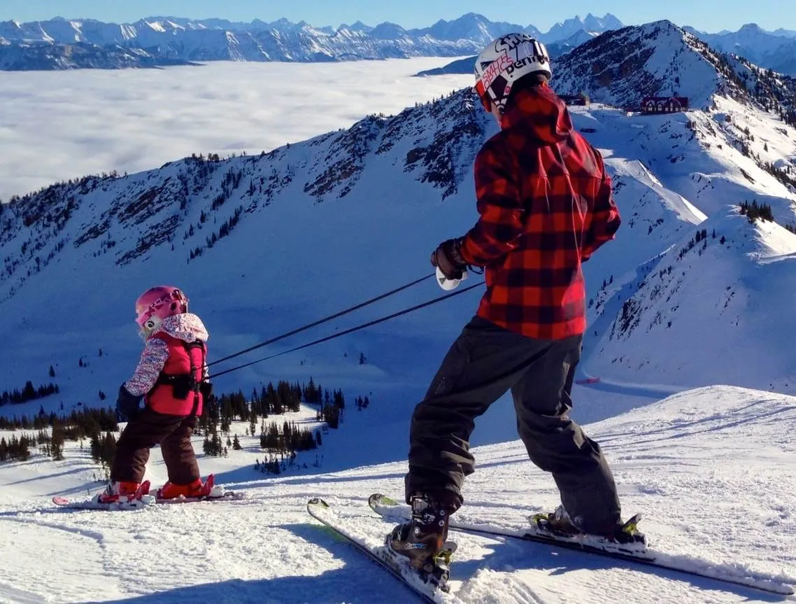 Father teaches child to ski with Lil' Ripper Gripper. The child wears a harness with leashes on a snow-covered mountain landscape.