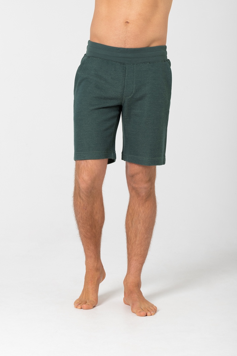Mann trägt grüne, gestrickte, kurze Hose mit Gummibund und Taschen, barfuß auf weißem Grund.