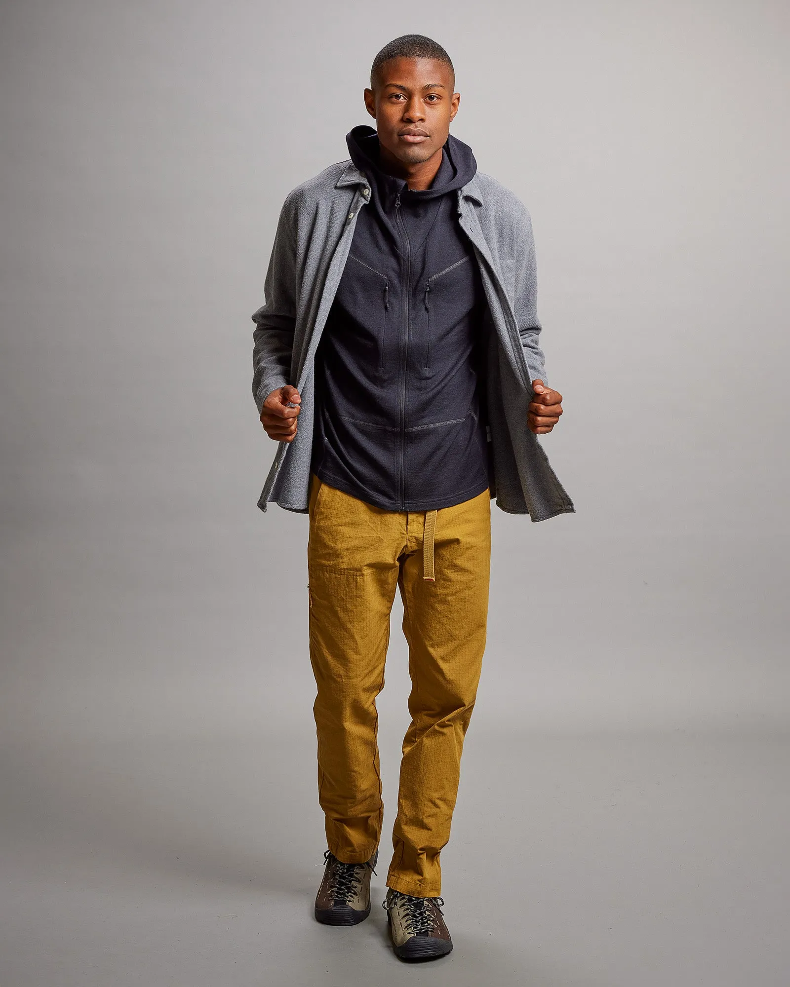 L'homme porte une veste à capuche Nuyarn noire sous une chemise grise, ainsi qu'un pantalon ocre et des chaussures de marche.