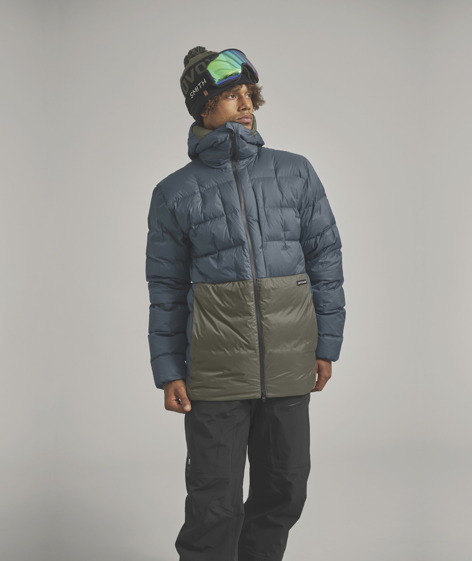 Ein Mann mit Skibrille und Mütze trägt eine zweifarbige Daunenjacke in Blau und Grün und schwarze Skihosen.