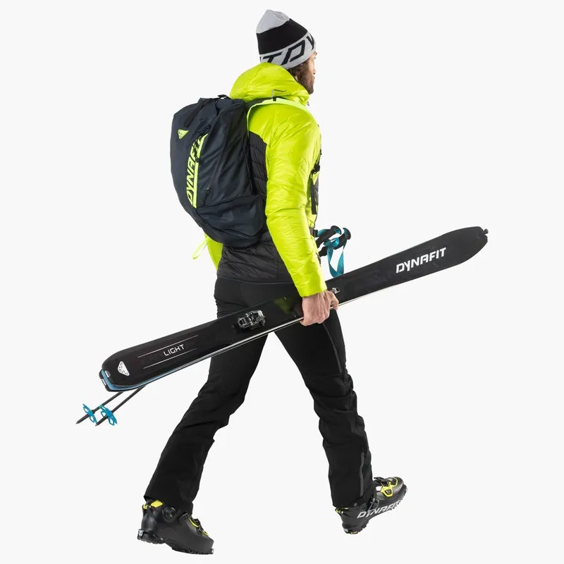 Ein Mann mit Rucksack und Dynafit Blacklight 95 Ski unter dem Arm, seitliche Ansicht.