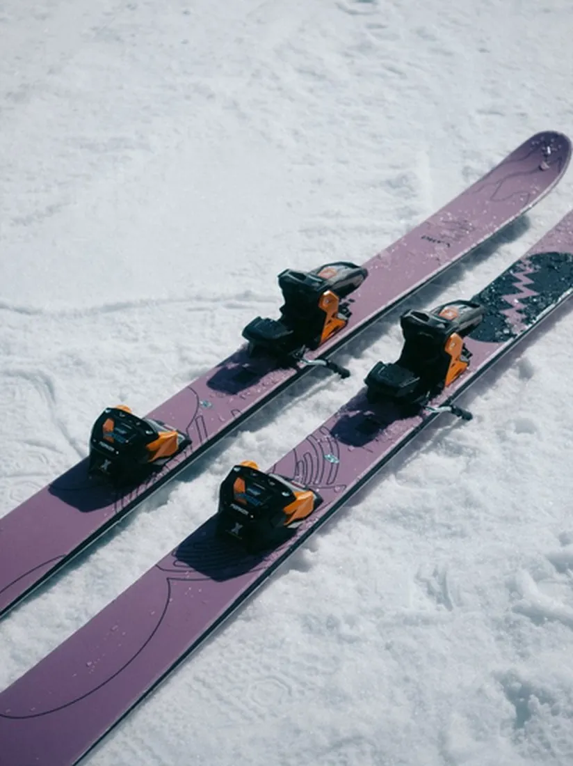 Due sci viola con attacchi arancioni e neri giacciono nella neve. Gli sci hanno un motivo grafico.