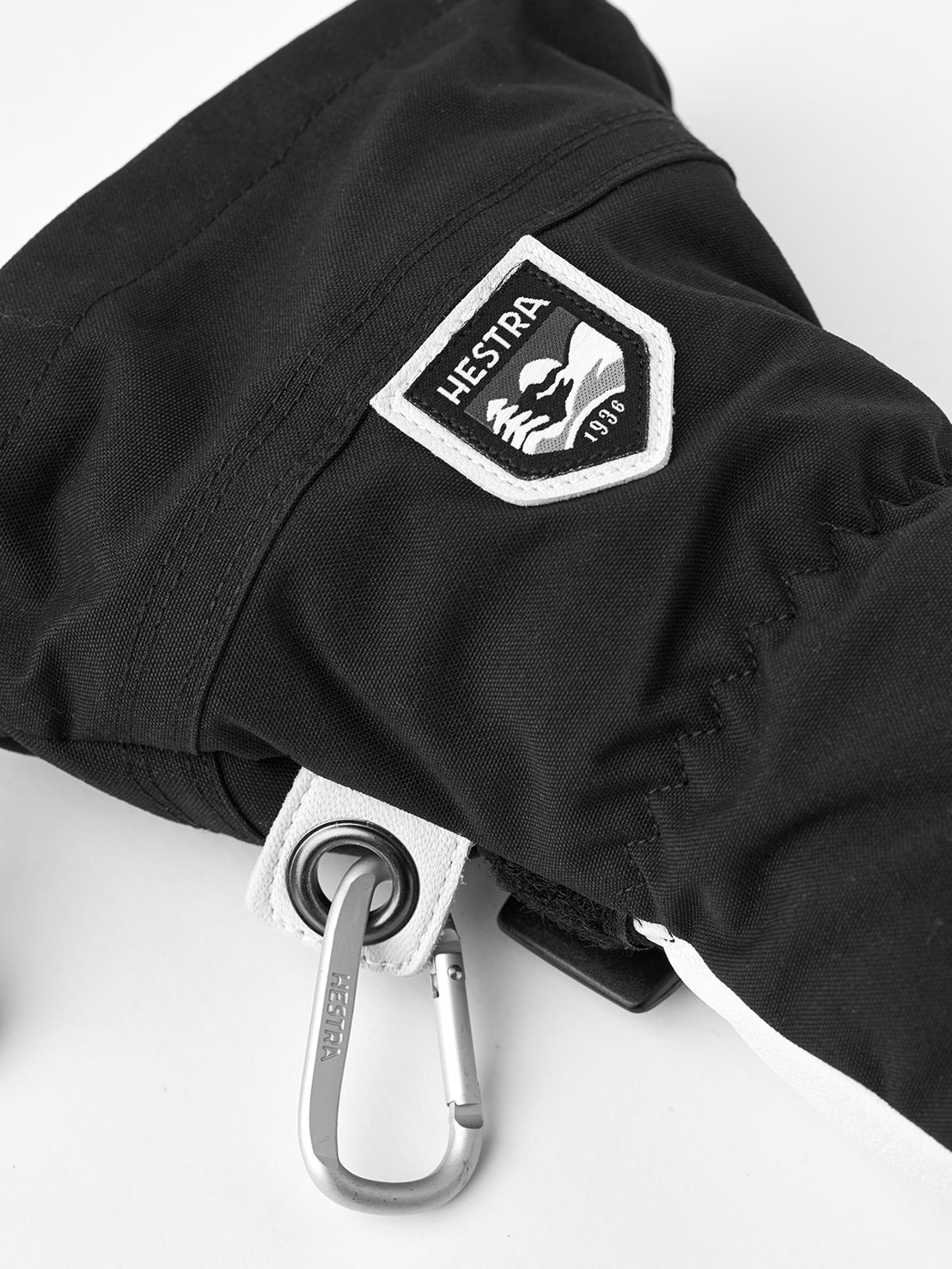 Detailaufnahme eines schwarzen Hestra Handschuhs mit Logo und Karabinerhaken.
