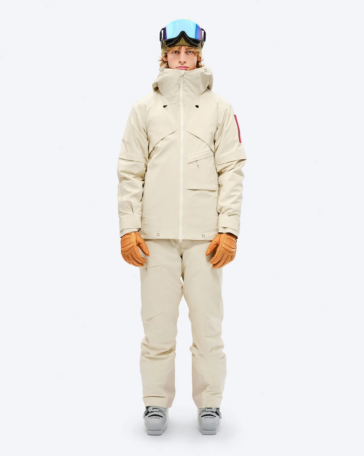 Person in beiger Ski-/Snowboardjacke mit GORE-TEX, Hose, Handschuhen und Skibrille vor weißem Hintergrund.