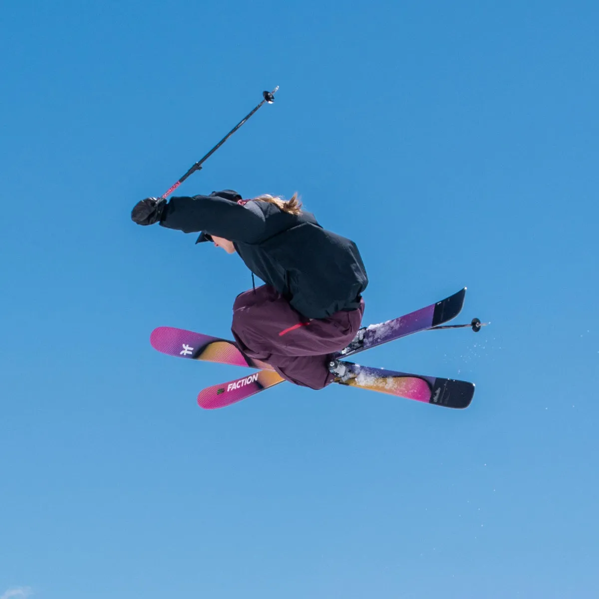 Skier in the air in front of a blue sky, with crossed skis and a ski pole in her hand.