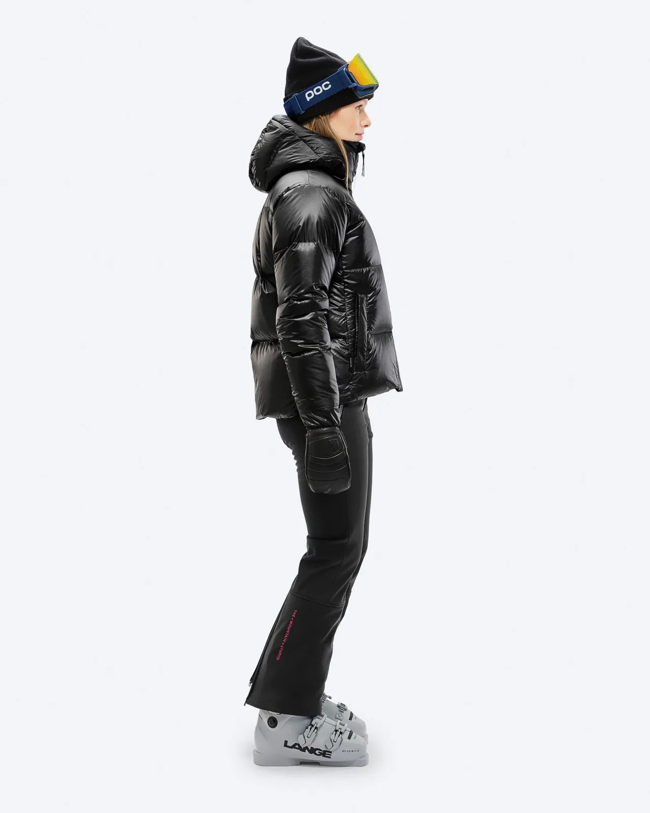 Frau in schwarzer Skihose und Jacke von der Seite, mit Skischuhen, Mütze und Skibrille.