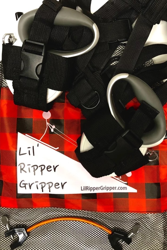 Lil' Ripper Gripper: Détail de l'aide à l'apprentissage du ski avec laisses, sangle, poignées et porte-pointes de ski sur tissu à carreaux rouge et noir.