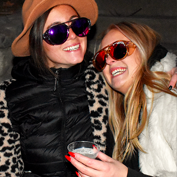 Due donne sorridenti con occhiali da sole polarizzati Smoked Show, in piedi vicine e con un bicchiere in mano.