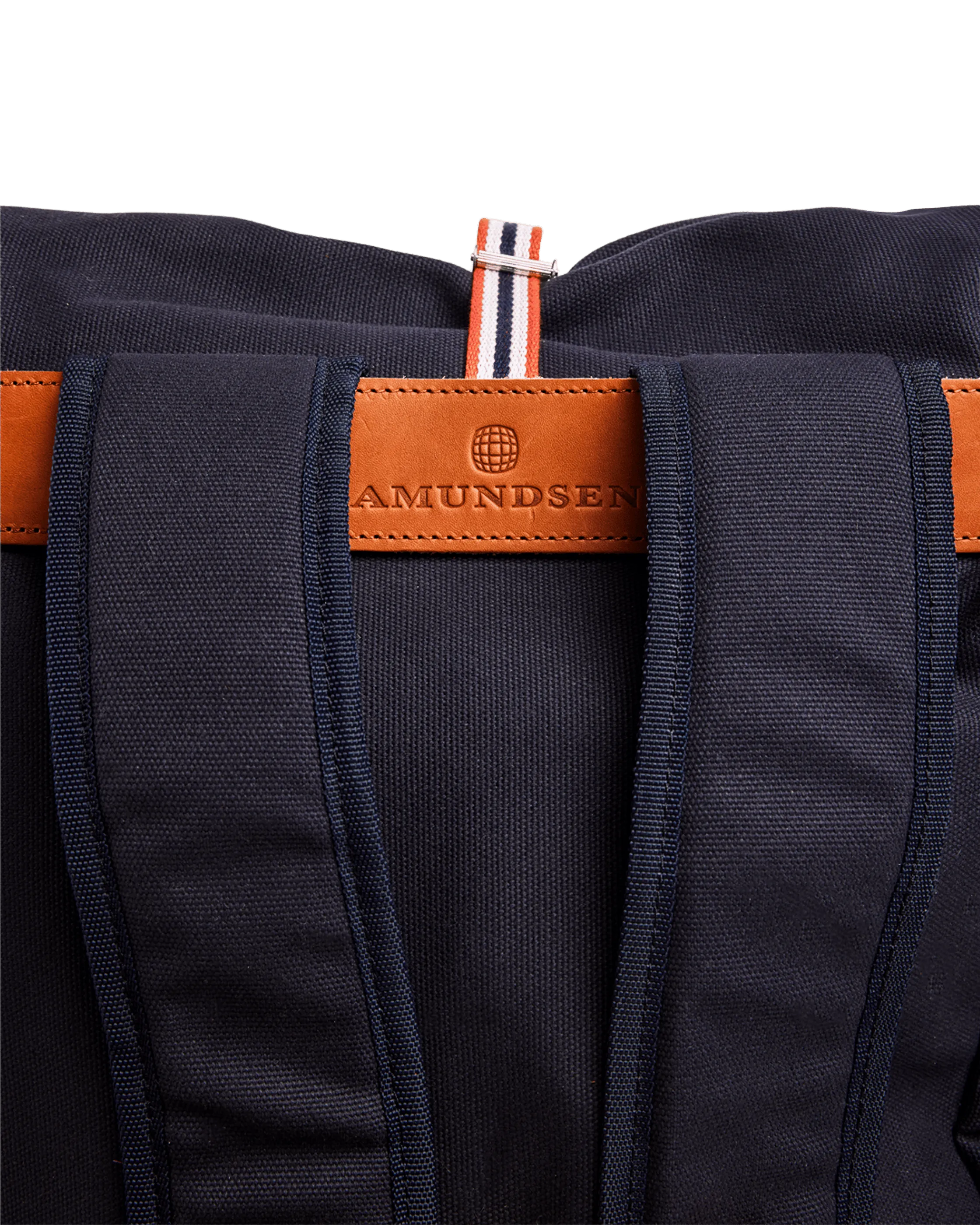 Primo piano di uno zaino blu scuro con patch in pelle marrone "Amundsen" e tracolla a righe arancioni, bianche e blu.