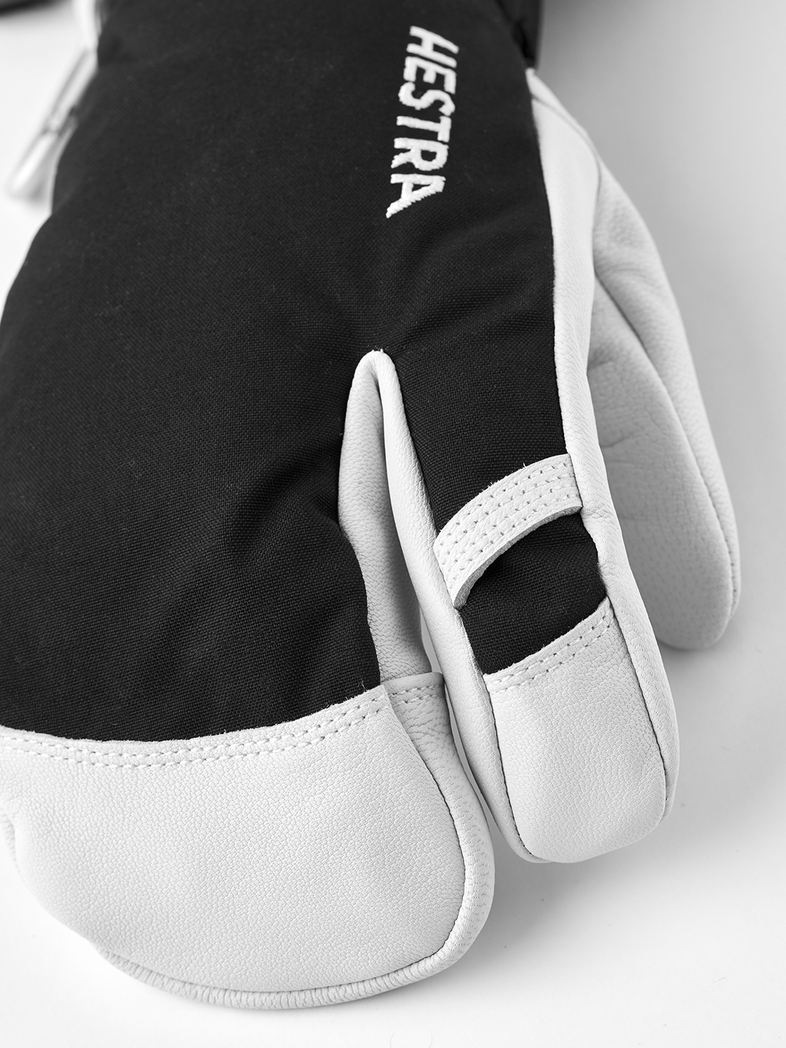 Hestra Gant à 3 doigts Gros plan sur un gant noir Hestra à 3 doigts avec du cuir blanc sur les doigts et le pouce. Le logo Hestra est visible verticalement.
