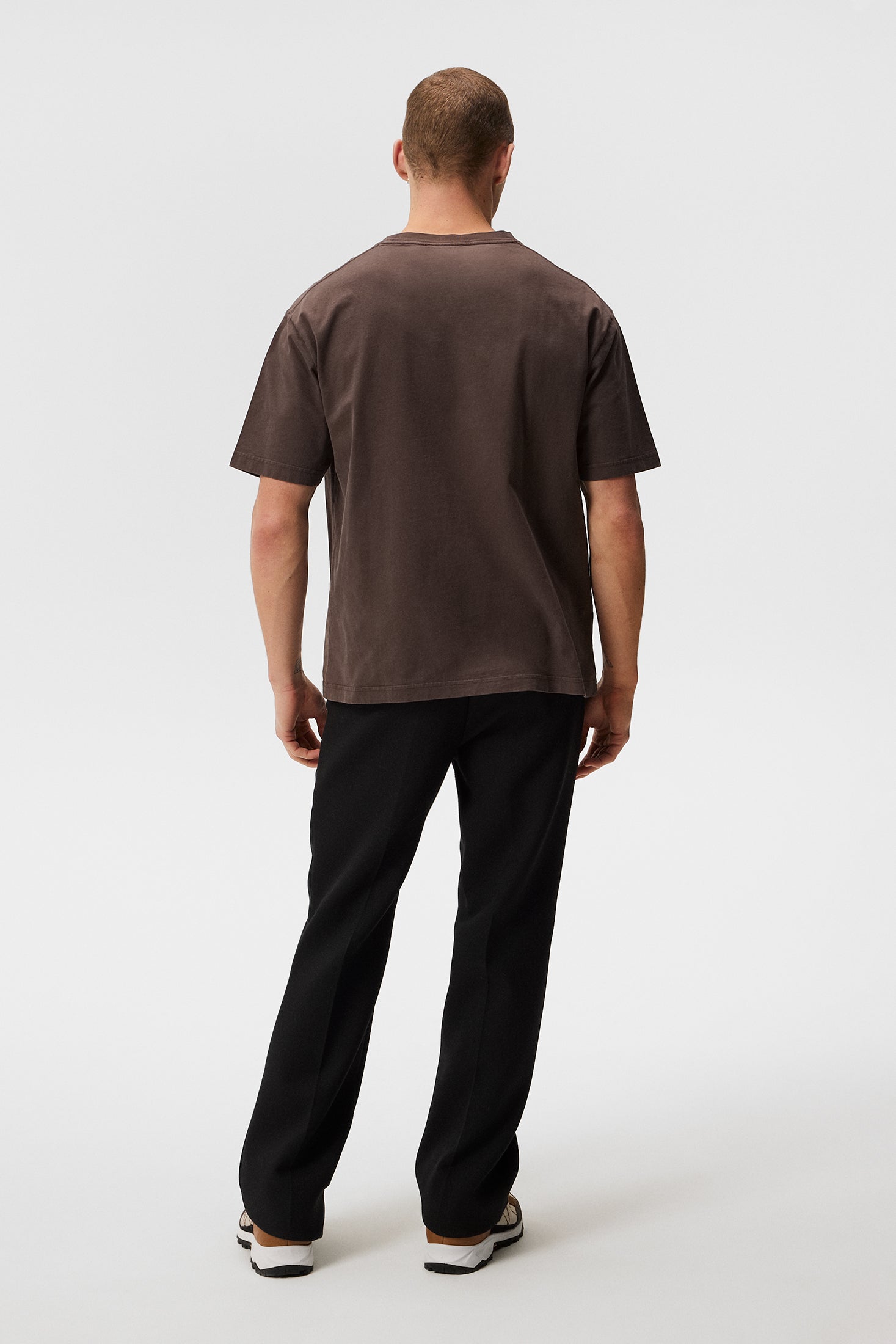 Ein Mann von hinten, trägt ein braunes T-Shirt und schwarze Hose vor einem weißen Hintergrund.