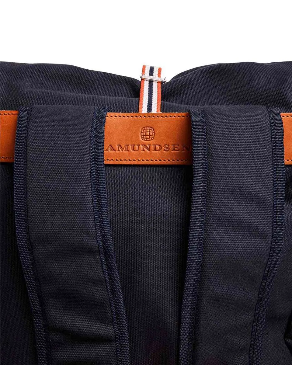 Primo piano di uno zaino blu scuro con patch in pelle marrone "Amundsen" e tracolla a righe arancioni, bianche e blu.