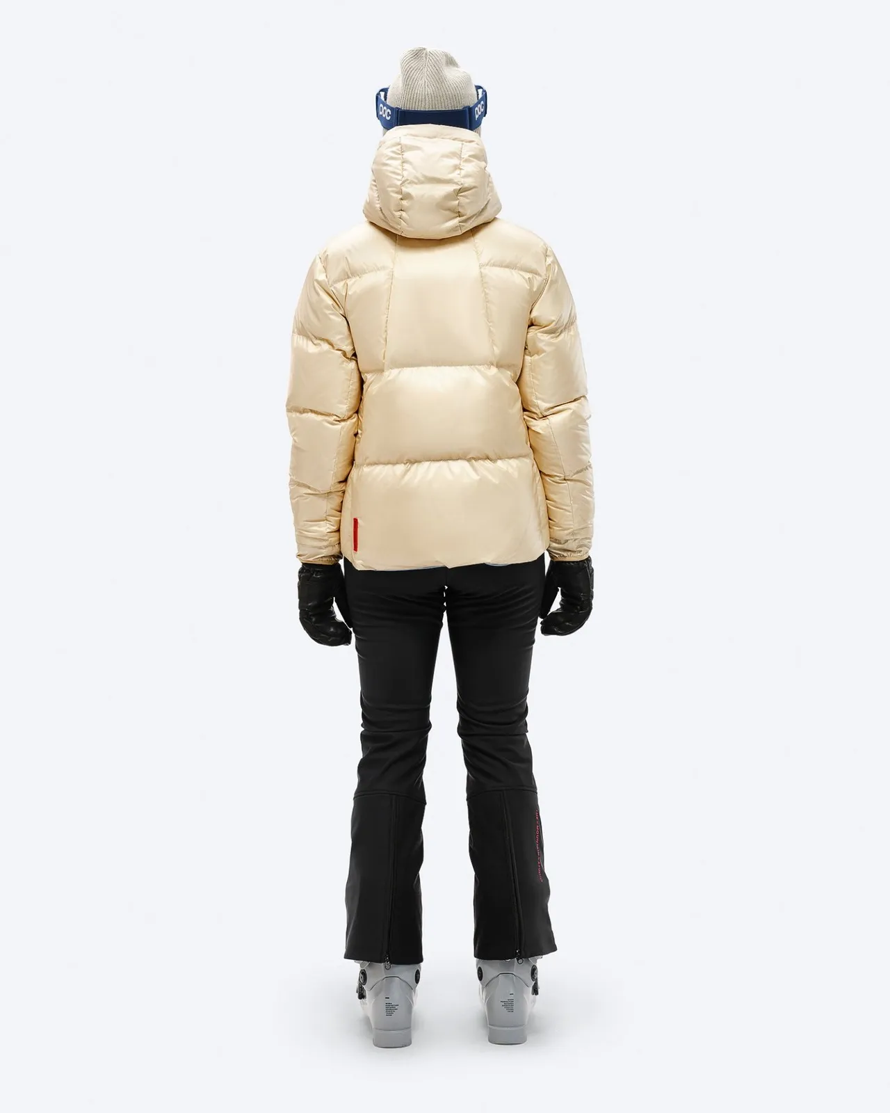 Back view: Beige down jacket with hood, black ski pants, gloves and ski boots, against a white background.