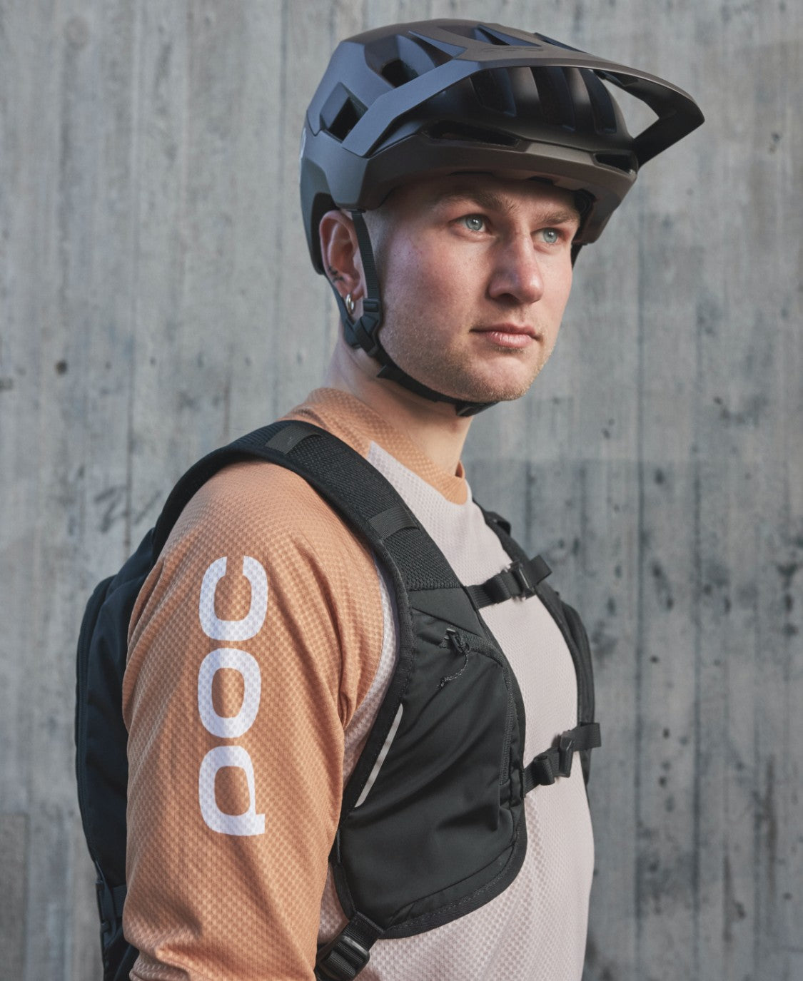 Mann mit VPD Rucksack Ein Mann mit Fahrradhelm und Column VPD Backpack 13L vor einer Betonwand. Er trägt ein langärmliges Hemd mit "POC" auf dem Ärmel.