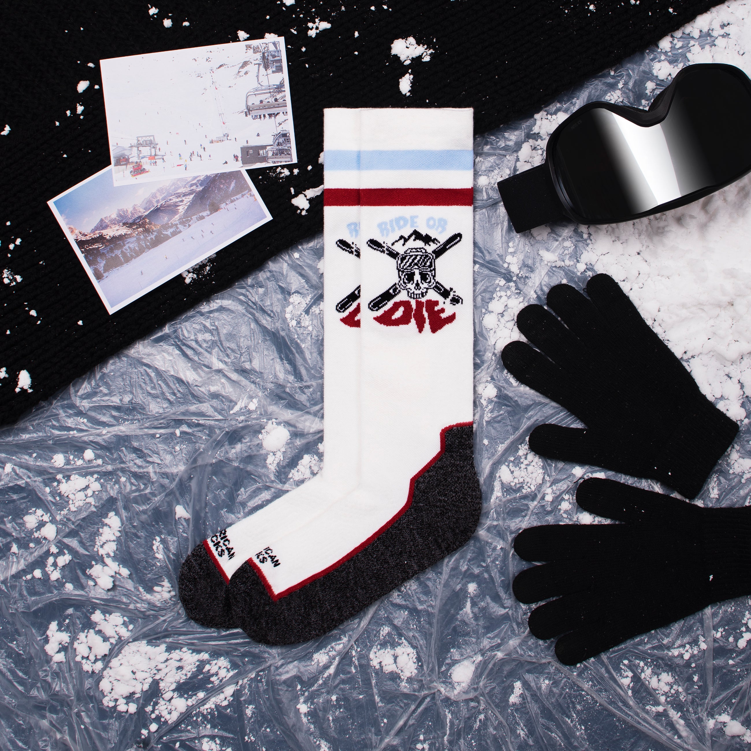 Ride or Die Kniestrümpfe Weiße Kniestrümpfe mit "Ride or Die" Motiv neben Schneebrille, Handschuhen, Mütze und Winterfotos auf vereister Oberfläche.