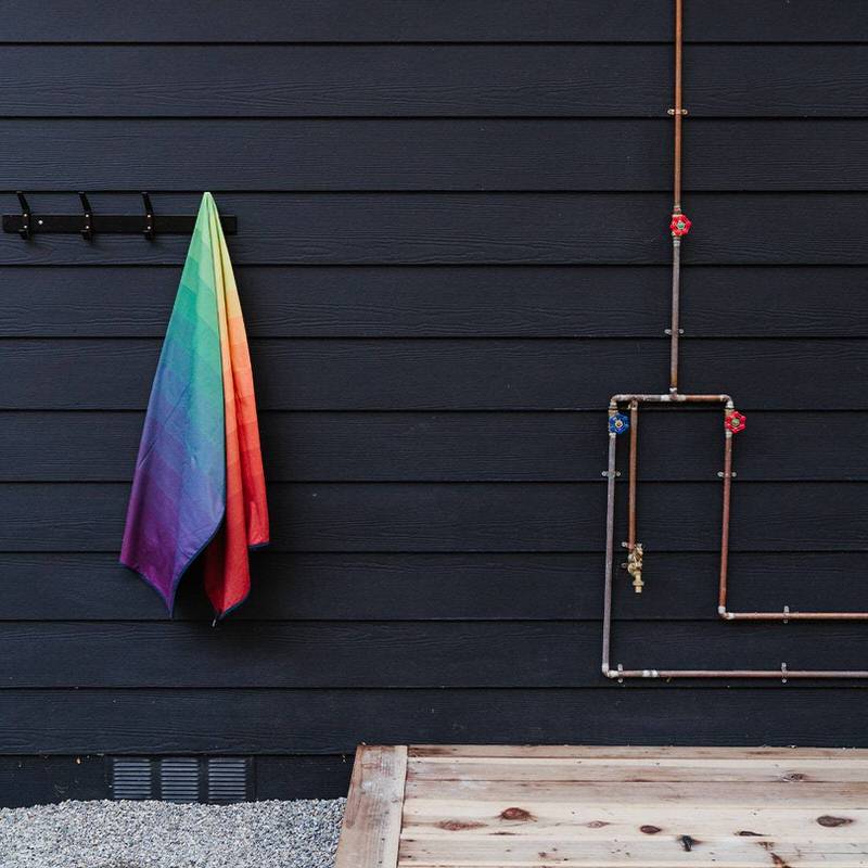 Regenbogenfarbenes Rumpl Shammy Towel hängt an Haken vor dunkler Holzfassade mit Kupferrohren.