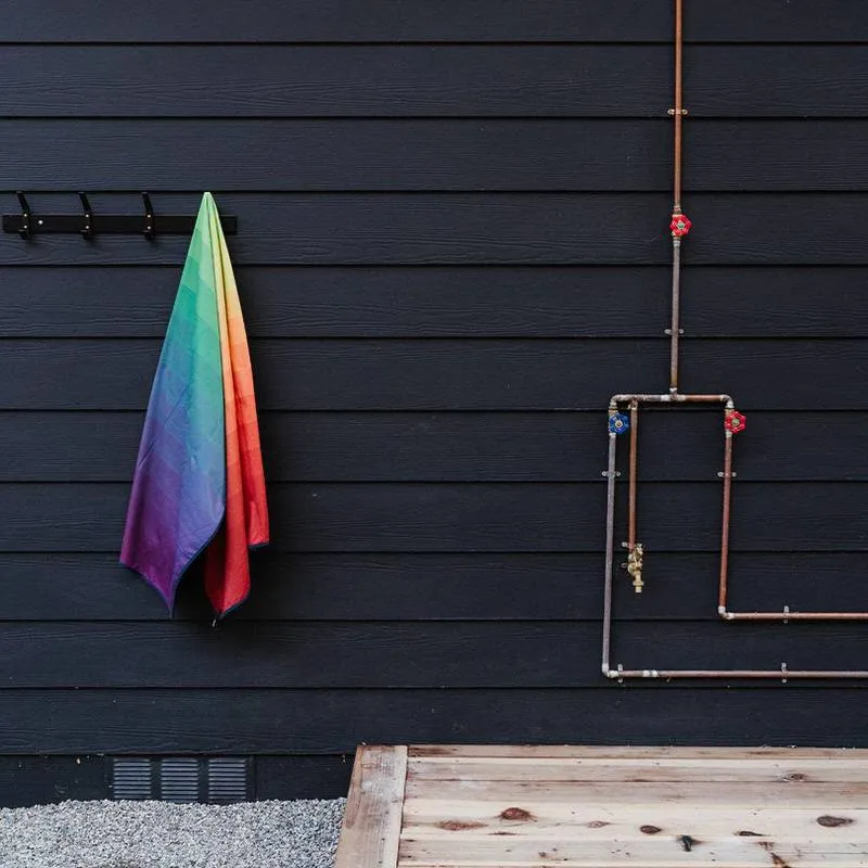 Regenbogenfarbenes Rumpl Shammy Towel hängt an Haken vor dunkler Holzfassade mit Kupferrohren.