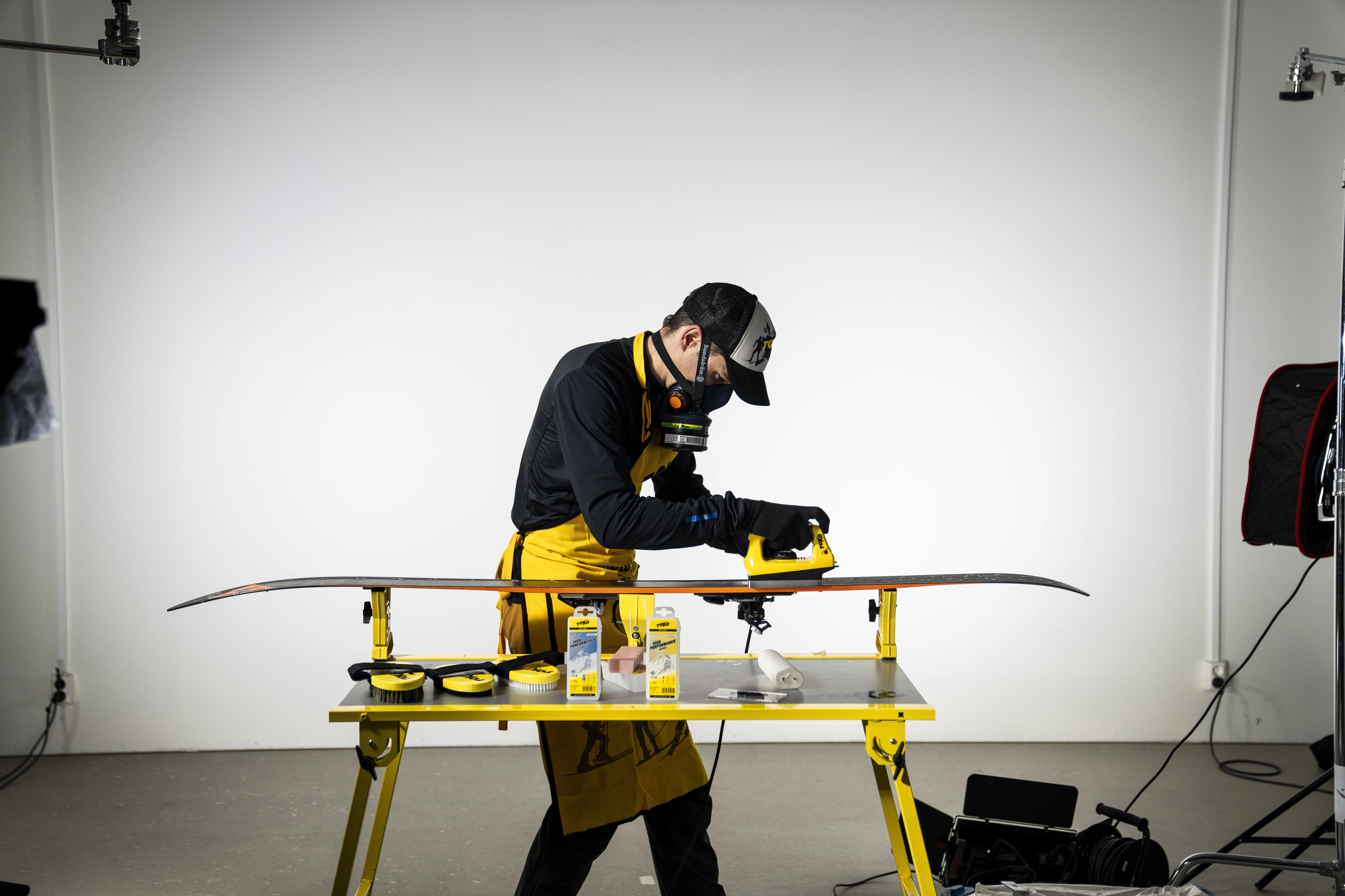 Man with mask and apron waxing skis on a yellow table. Tools and accessories are arranged on it, background is bright.
