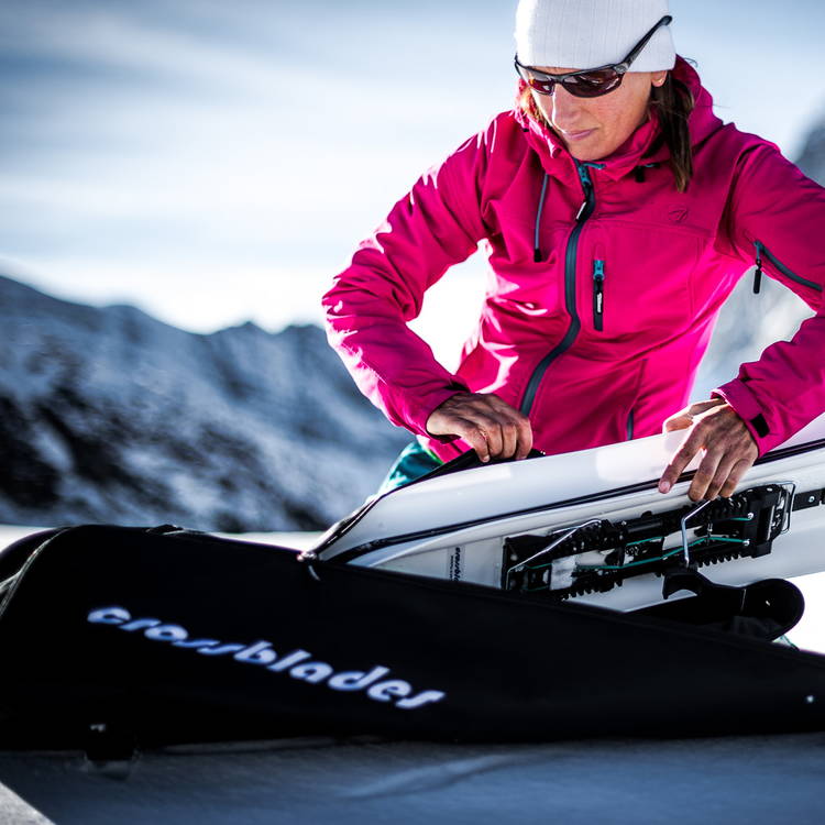 Frau packt Crossblades in eine schwarze Tasche mit Logo vor schneebedeckter Bergkulisse.
