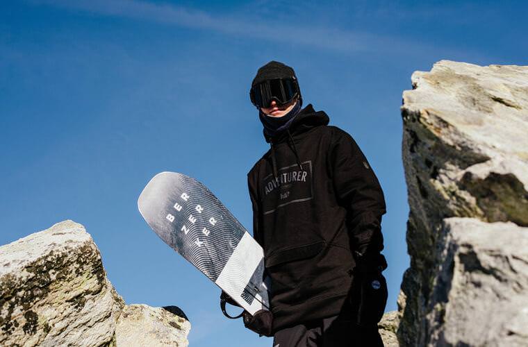 Person mit Navy Neckwarmer, Snowboard, Mütze und Brille vor blauem Himmel.