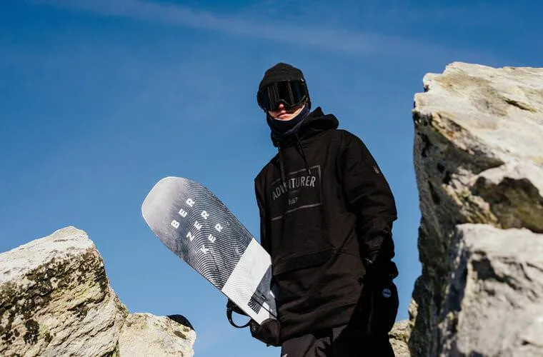 Person mit Navy Neckwarmer, Snowboard, Mütze und Brille vor blauem Himmel.