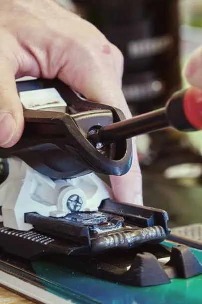 One hand adjusts the ski binding on a ski with a screwdriver. The binding is black and white.