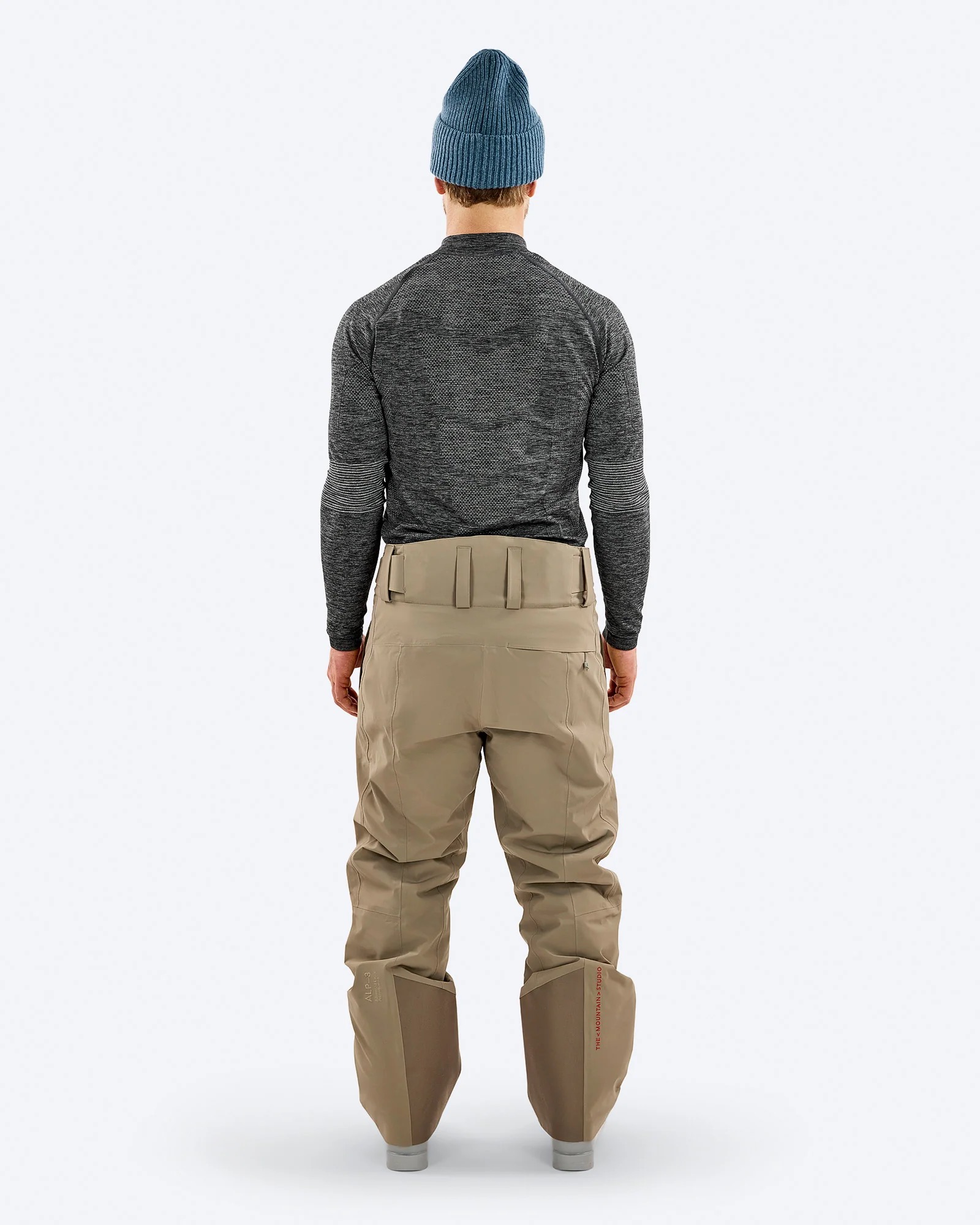 Ein Mann von hinten mit blauer Mütze, grauem Langarmshirt und beigen Skihosen auf weißem Hintergrund.