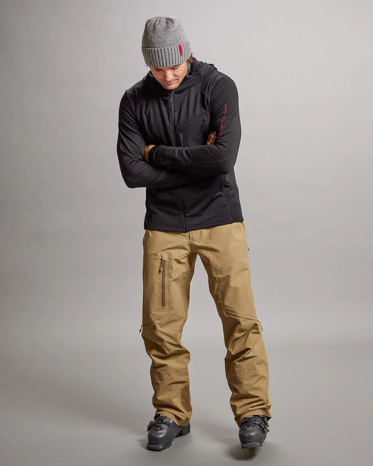Man in black M-4 Techno Stretch Mid Hood, beige pants and gray cap, posing with folded arms.