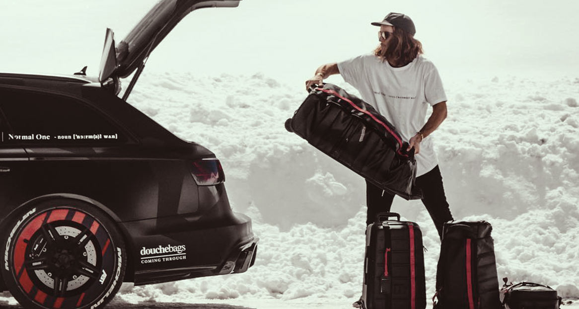 Ein Mann packt Taschen aus einem schwarzen Kombi mit geöffneter Heckklappe in einer verschneiten Landschaft.
