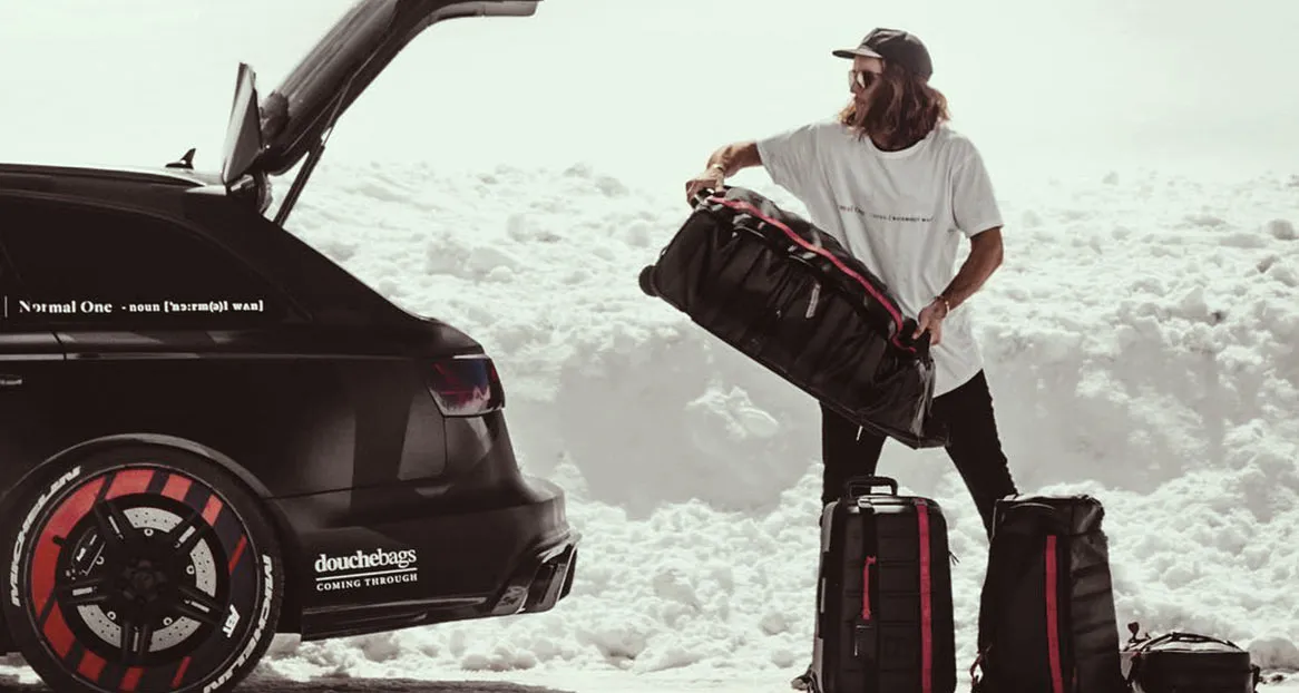 Ein Mann packt Taschen aus einem schwarzen Kombi mit geöffneter Heckklappe in einer verschneiten Landschaft.