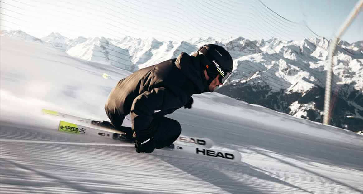 Ein Skifahrer in schwarzer Jacke, Hose und Helm fährt eine präparierte Piste hinunter, im Hintergrund verschneite Berge.