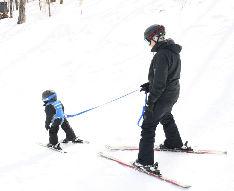 An adult teaches a small child to ski. The child wears a blue harness and is guided by a leash.