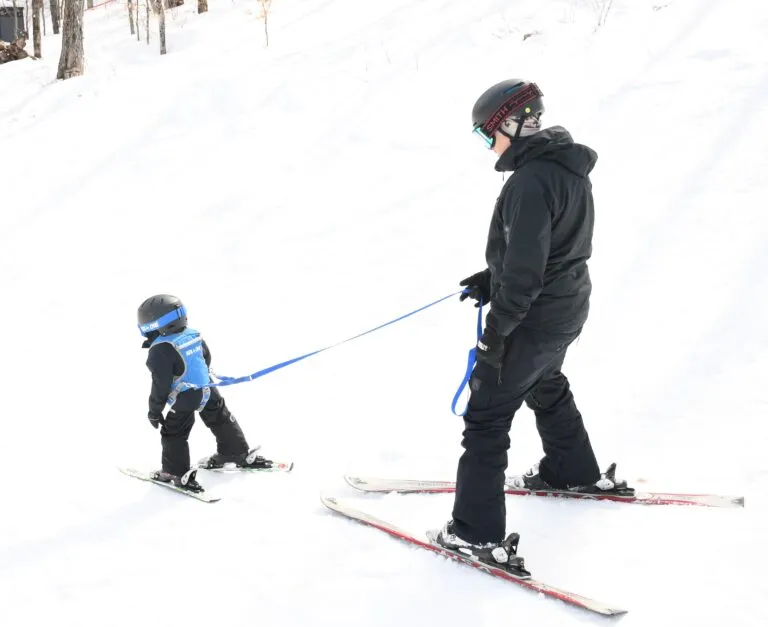 An adult teaches a small child to ski. The child wears a blue harness and is guided by a leash.