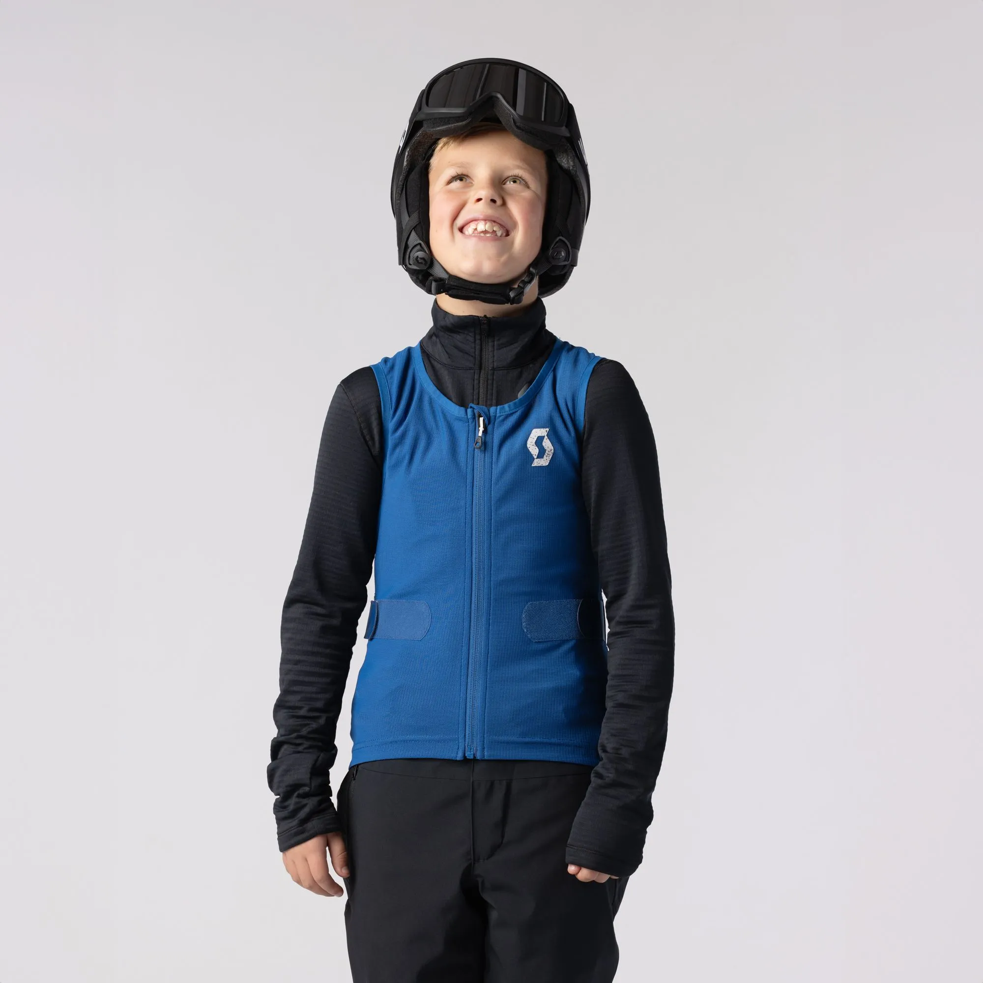Ein lächelnder Junge mit Helm und Skibrille trägt eine blaue SCOTT Airflow Junior-Weste.