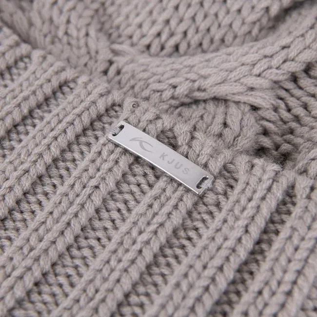Gros plan sur un bonnet gris clair en tricot avec un motif torsadé et une étiquette argentée avec le logo "KJUS".