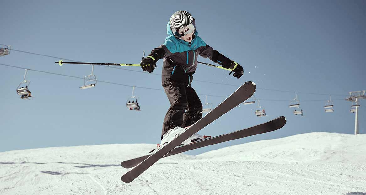 Ein Kind springt auf Skiern über eine schneebedeckte Piste. Im Hintergrund ist ein blauer Himmel und eine Seilbahn zu sehen.