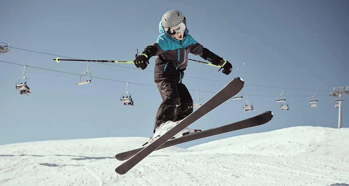 Ein Kind springt auf Skiern über eine schneebedeckte Piste. Im Hintergrund ist ein blauer Himmel und eine Seilbahn zu sehen.