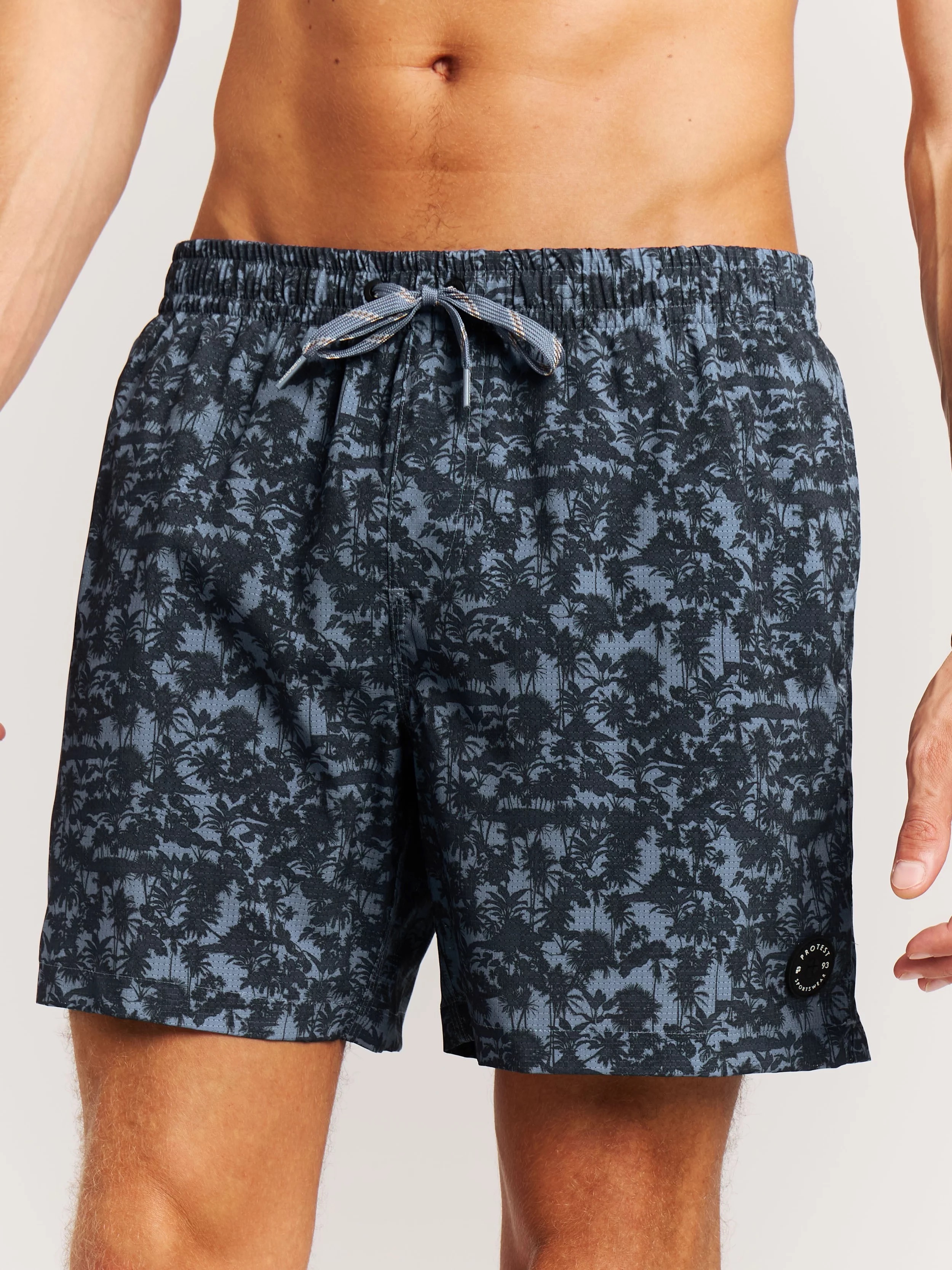 Ein Mann trägt Badehosen mit einem schwarzen Palmenmuster auf blauem Grund. Die Badehose hat eine Kordel am Bund.