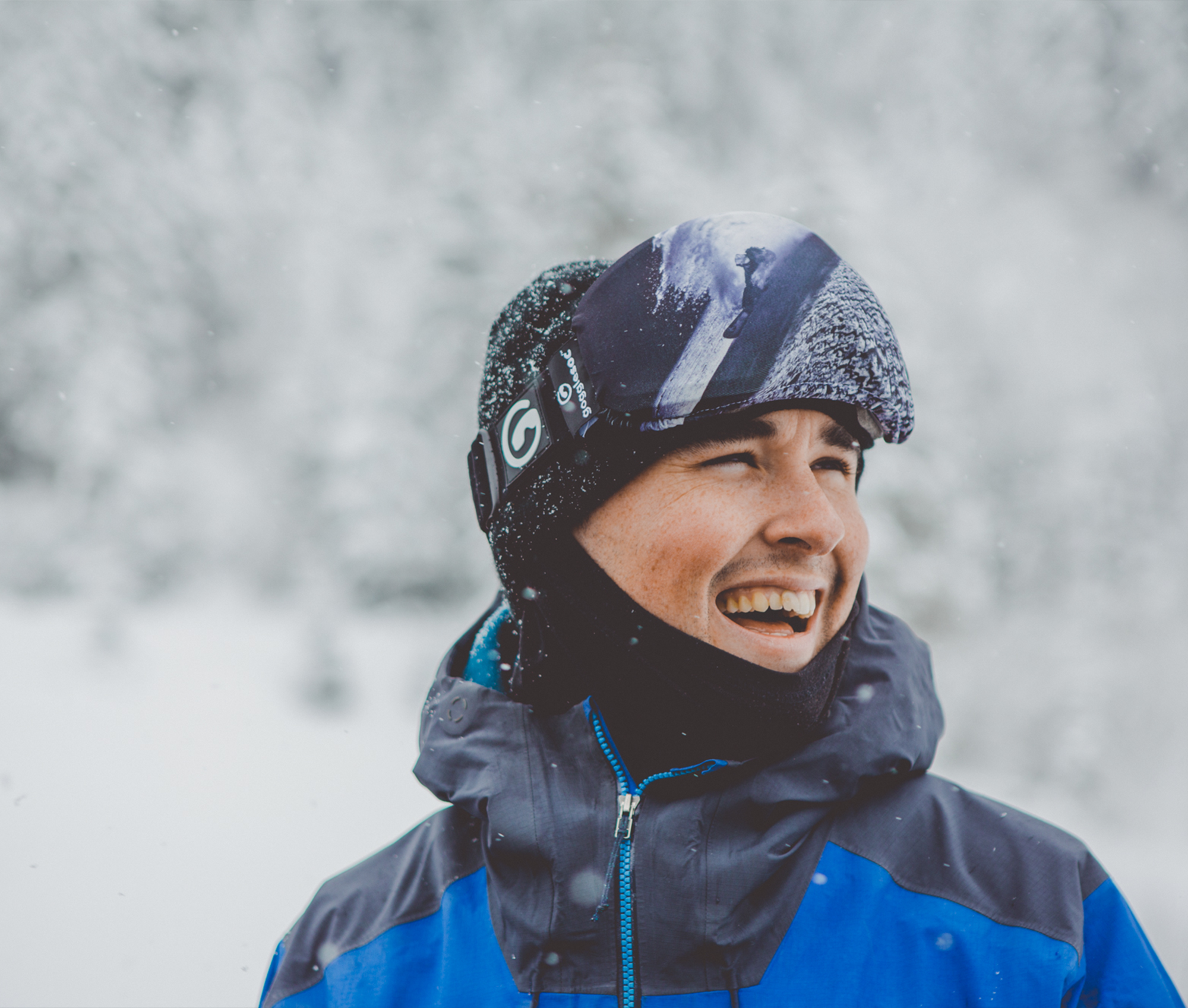 Lächelnder Mann mit Skibrille und Mütze im Schnee, trägt eine blau-graue Jacke, Teil der "Moon Dust Soc" Kollektion.