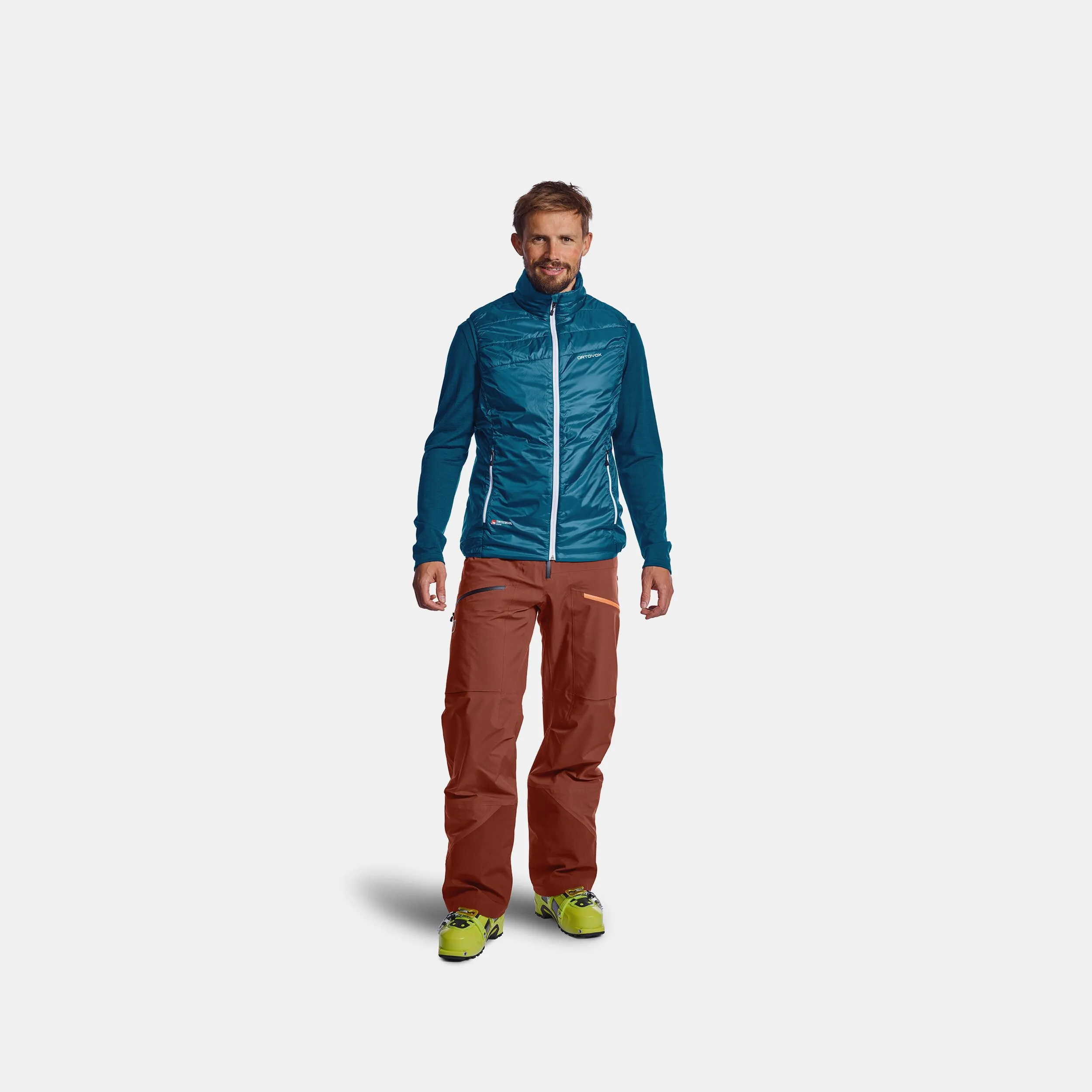 A man wears the blue Ortovox PIZ Cartas SWISSWOOL Vest with zipper, a long blue jacket and ski pants.