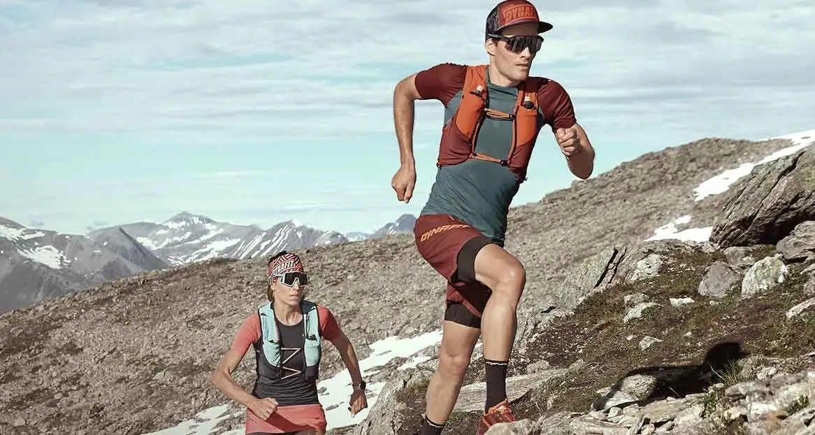 Two trail runners in sportswear run uphill. Snow-covered mountains and a cloudy sky are visible in the background.