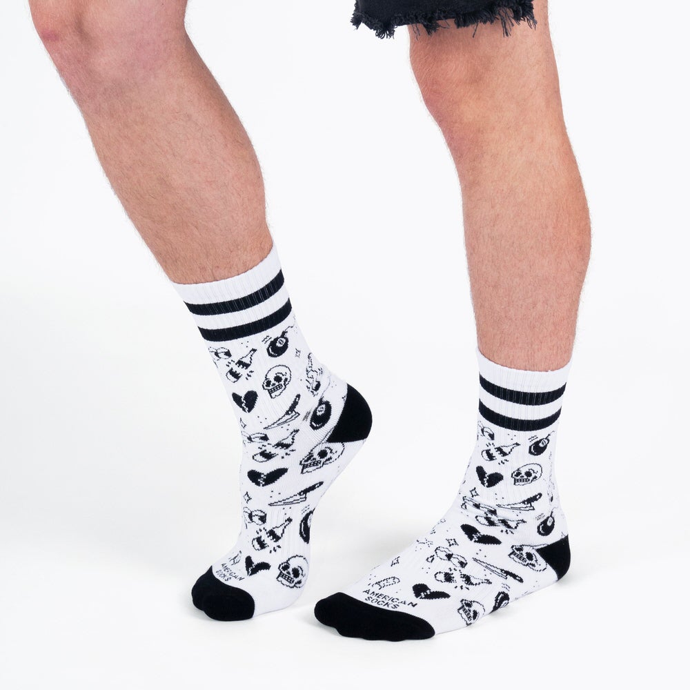 Beine mit weißen, mittelhohen Socken, schwarze Bündchen und Streifen. Socken mit Flash Tattoo Motiven, wie Totenköpfe.